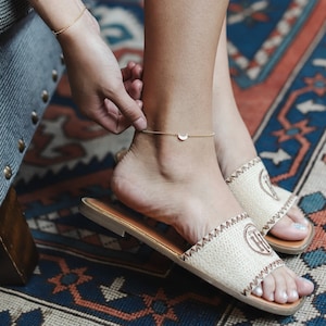 May include: A gold anklet with a crescent moon charm. The anklet is worn on a person's ankle, and the person is wearing beige sandals.