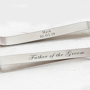 May include: Two silver tie clips with engraved text. The top clip reads "Mark 10.05.19". The bottom clip reads "Father of the Groom".