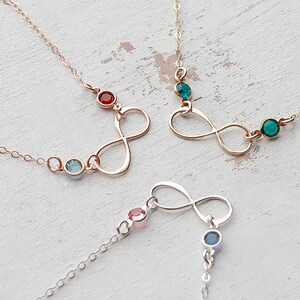 May include: Three infinity symbol necklaces with different colored gemstones. The necklaces are on a white wooden surface. The gemstones are red, blue, and green.