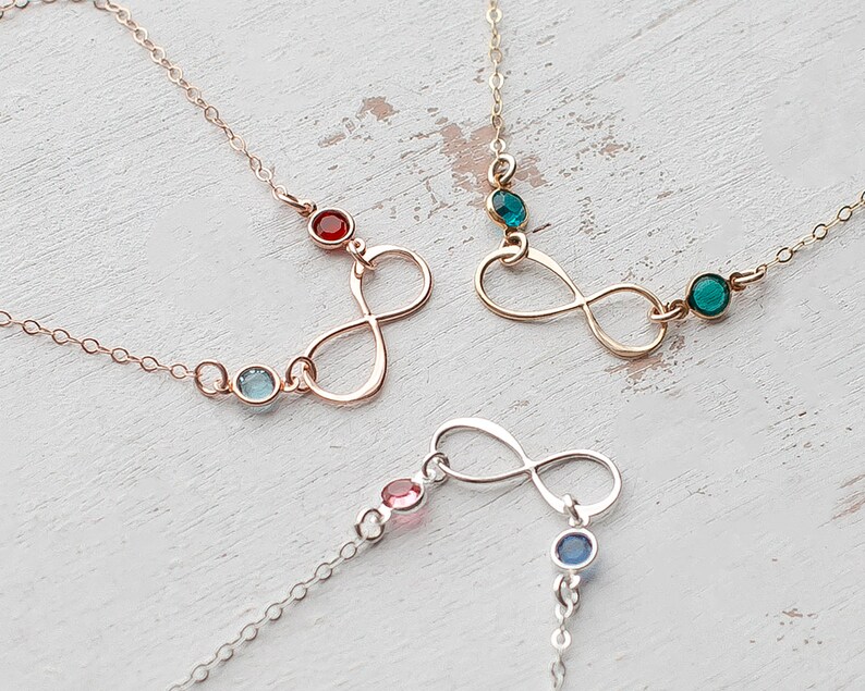 May include: Three infinity symbol necklaces with different colored gemstones. The necklaces are on a white wooden surface. The gemstones are red, blue, and green.