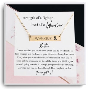 May include: A gold necklace with a bar pendant that reads "WARRIOR" with a pink ribbon charm. The necklace is in a box with a pink and white marble pattern and a message that reads "strength of a fighter heart of a Warrior" and "Cancer teaches you to treasure every day, to love freely, to find courage and to discover your faith even during hard times. Every time you wear this necklace remember what you've been able to overcome so far. All the times you felt like you weren't going to make it through, you proved yourself wrong. Warriors like you are born through life's toughest battles. You've got this!"