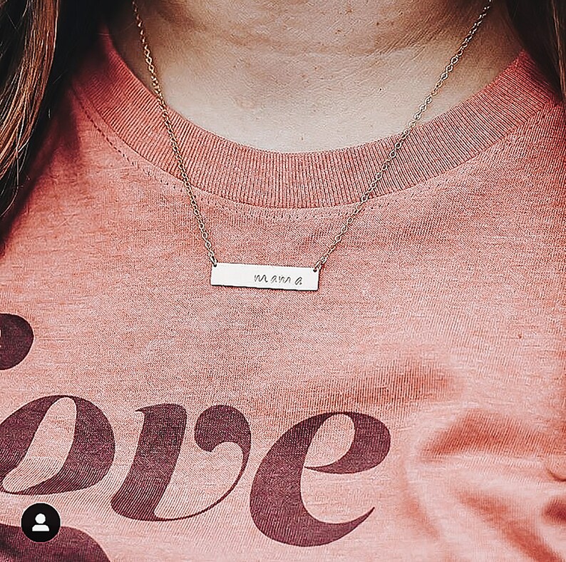 May include: A gold necklace with a bar pendant that says "mama" on it. The necklace is worn by a person wearing a pink shirt with the word "love" printed on it.