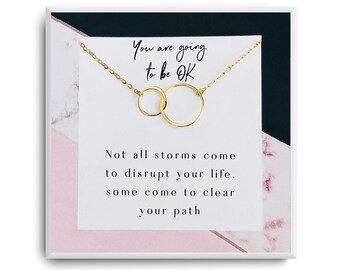 May include: A gold necklace with two interlocking circles, packaged in a white box with a pink and black marble design. The box includes a card with the text "You are going to be OK. Not all storms come to disrupt your life, some come to clear your path."