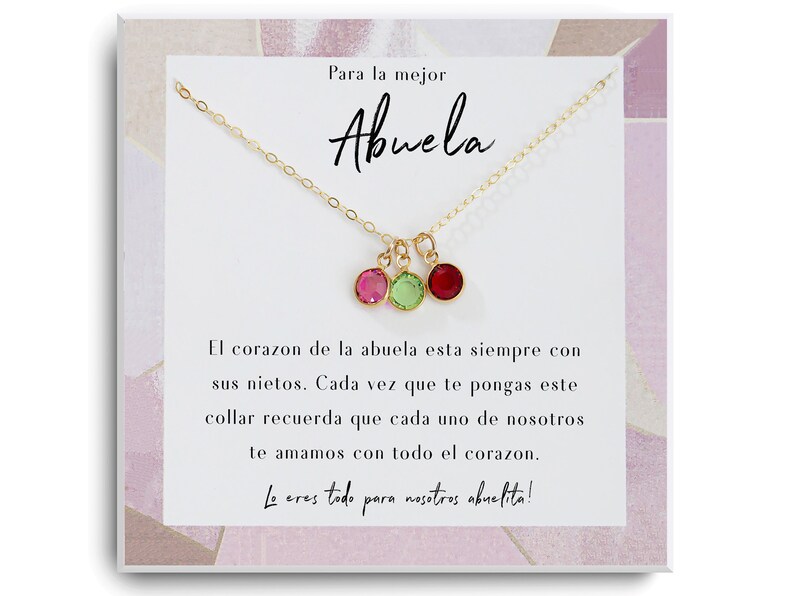 May include: A gold necklace with three small colorful gemstones hanging from it. The necklace is displayed on a card with the text "Para la mejor Abuela" and a message in Spanish about the love of a grandmother.