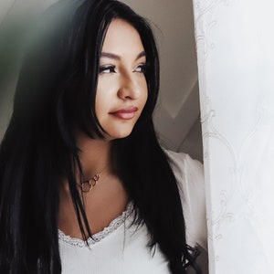 May include: A woman with long black hair wearing a white top with a lace trim. She is wearing a gold necklace with multiple circles. She is looking to the right of the frame.