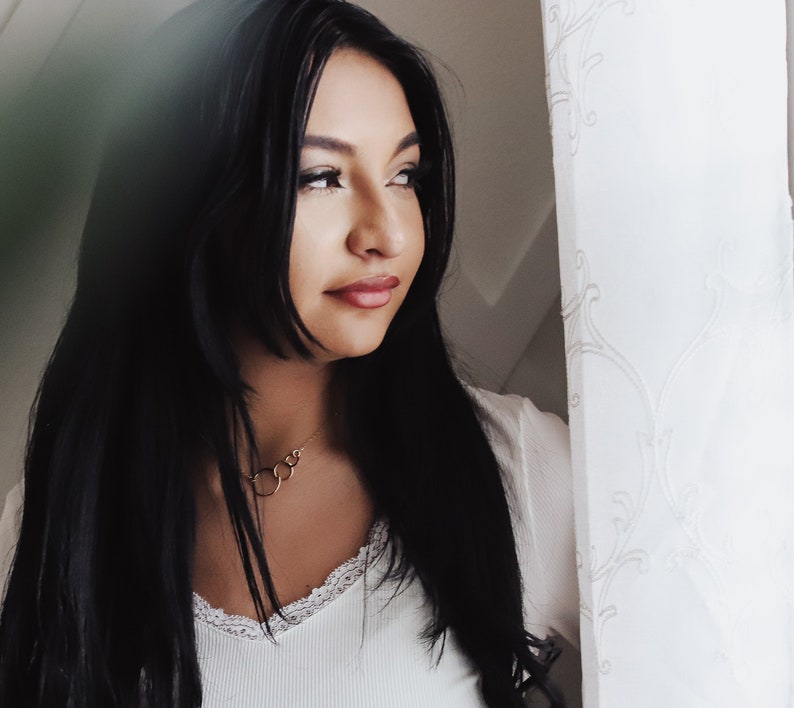 May include: A woman with long black hair wearing a white top with a lace trim. She is wearing a gold necklace with multiple circles. She is looking to the right of the frame.