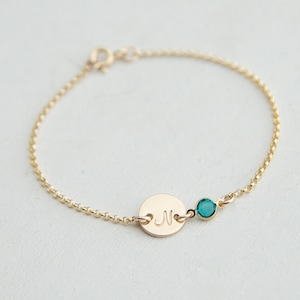 May include: A gold chain bracelet with a small round charm engraved with the letter "N" and a teal blue gemstone.
