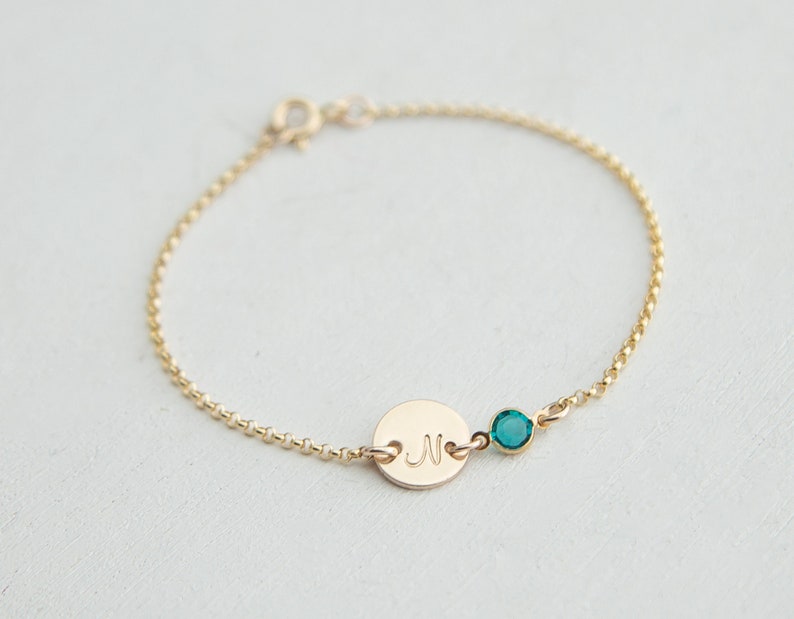 May include: A gold chain bracelet with a small round charm engraved with the letter "N" and a teal blue gemstone.
