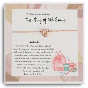 Op de afbeelding: Een roze kaart met een boodschap om iemand een fijne eerste dag in groep 4 te wensen. De kaart heeft een roze rugzak, boeken en een kopje koffie erop. Er is een roségouden ketting met een hartvormige hanger met de letter M erop.