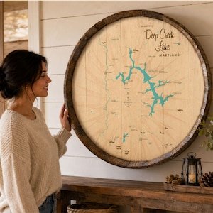 May include: A circular wall art piece crafted from a wooden barrel head, featuring a map of Deep Creek Lake, Maryland. The map displays the lake in teal blue against a light wood grain background, with black text labels and a compass rose.