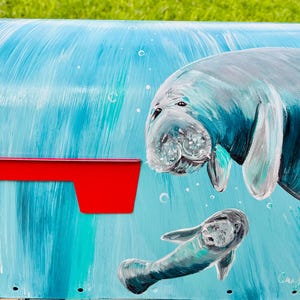May include: A mailbox painted with a light blue background and a red flag. The mailbox features a hand-painted image of a manatee and its calf. The manatees are painted in shades of gray and blue.