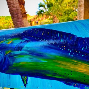 May include: A vibrant painting of a mahi-mahi fish in shades of blue, green, and yellow. The artwork depicts the fish swimming in a stylized ocean, with brushstrokes creating a sense of movement. The background is a bright blue.