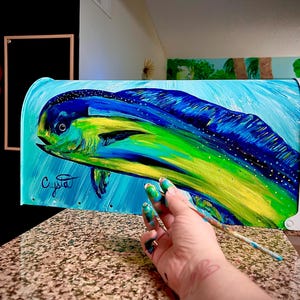 May include: A mailbox painted with a vibrant depiction of a mahi-mahi fish. The artwork features shades of blue, green, and yellow, with the artist's signature visible. The mailbox is set against a light blue background.