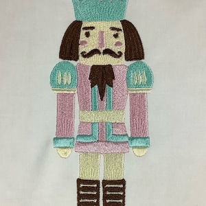 May include: A pink and teal embroidered nutcracker with a brown beard and hat. The nutcracker is standing on a white background. Design 5.