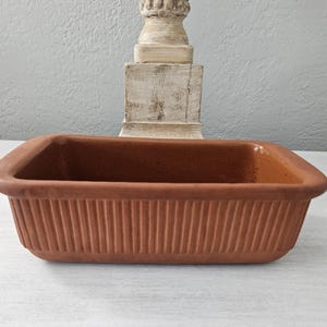 May include: A rectangular terracotta baking dish with a ribbed exterior texture and a smooth interior finish. The vessel features a wide, flat rim and deep walls, showcasing the natural reddish-brown clay color throughout the entire piece.