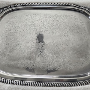 May include: A rectangular silver-toned serving tray with rounded corners and two ornate handles. The tray features a polished surface with an intricate engraved floral and scrollwork pattern in the center and a twisted rope-style border.