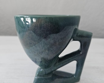 May include: A green ceramic cup with a unique, angular handle and a triangular base. The cup has a glossy finish and a subtle iridescent effect.