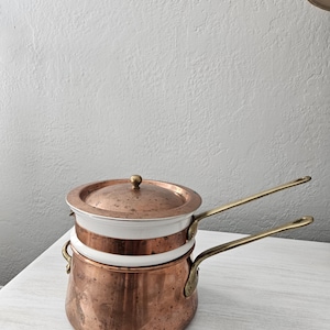 May include: A double boiler set featuring a copper base pot and a copper lid, both with a white enamel interior band. The set includes two long brass handles attached to the sides and a small brass knob on the center of the lid.