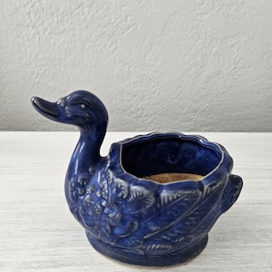 May include: A blue ceramic planter shaped like a duck. The duck has a detailed design of feathers and a small opening for planting.