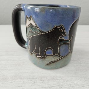 May include: A ceramic mug with a dark brown handle and a blue glaze finish. The exterior features a hand-carved design of a black bear standing in front of stylized mountain peaks. The base of the mug transitions into a lighter, muted blue tone.