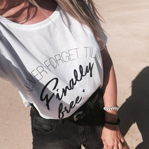 Puede incluir: Camiseta blanca con texto negro que dice "Never forget till finally free". La persona lleva pantalones cortos de mezclilla negros y una pulsera a cuadros blanco y negro.