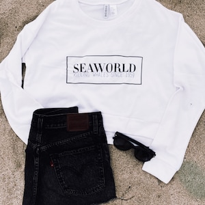 Puede incluir: Sudadera blanca con texto negro que dice "SEAWORLD KILLING WHALES SINCE 1959" en un cuadro. También se muestran pantalones cortos de mezclilla negros y gafas de sol negras.