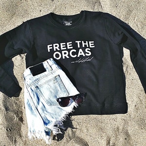 Puede incluir: Sudadera negra con el texto "FREE THE ORCAS" en letras blancas. Unos pantalones cortos de mezclilla azul claro con bordes deshilachados están encima de la sudadera.