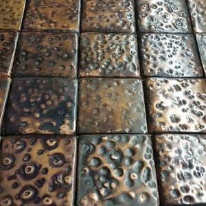 Op de afbeelding: Close-up van een koperen tegelvloer met een gehamerde textuur. De tegels zijn in een rasterpatroon gerangschikt en hebben een warme, metallic glans.