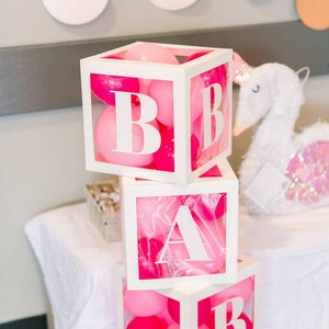 Op de afbeelding: Een stapel van vier witte houten blokken met de letters "BABY", elk gevuld met roze en felroze ballonnen. Een pluchen zwaan met roze pailletten staat in de buurt, wat wijst op een babyshower- of kinderkamerthema.