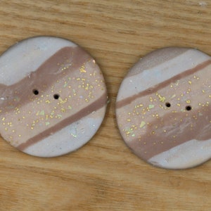May include: Two round, white buttons with brown stripes and gold glitter. The buttons have two holes.
