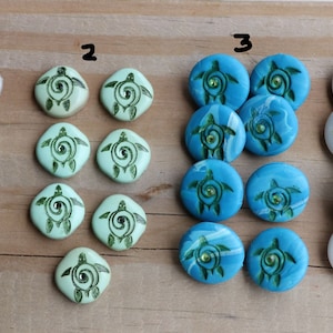 May include: A collection of handmade ceramic turtle-themed beads in various colors. The beads feature a turtle design with green accents and a central gem. Colors include white, light green, and blue.