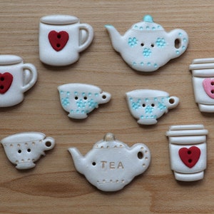 May include: A set of nine white ceramic buttons shaped like teacups, teapots, and mugs. The buttons have red and pink hearts and blue and white floral designs. The teapot button has the word "TEA" written on it.