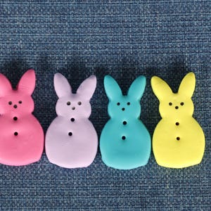 May include: Four colorful marshmallow bunny candies in pink, lavender, teal, and yellow. Each bunny has three black dot eyes and three black dot buttons. The bunnies are arranged on a blue textured surface.