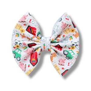 May include: A white fabric hair bow with a repeating pattern of farm animals, tractors, and red barns. The bow is tied in the center and has a textured appearance.