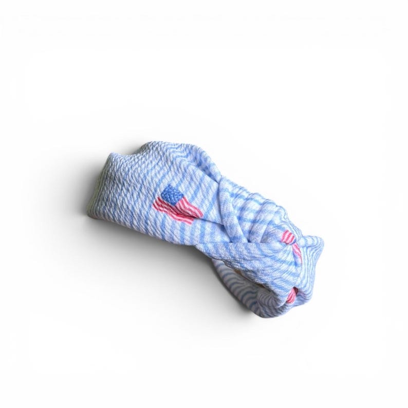May include: A blue and white striped headband with an embroidered American flag. The headband is twisted and knotted, with a soft, textured appearance. The flag features red and blue colors.