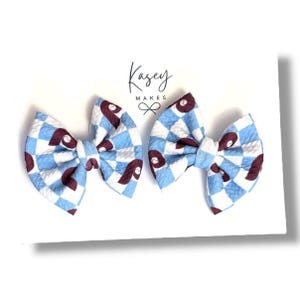 May include: Two blue and white checkered hair bows with maroon baseballs. The bows are tied with a white ribbon and have a "Kasey Makes" logo on the white background.