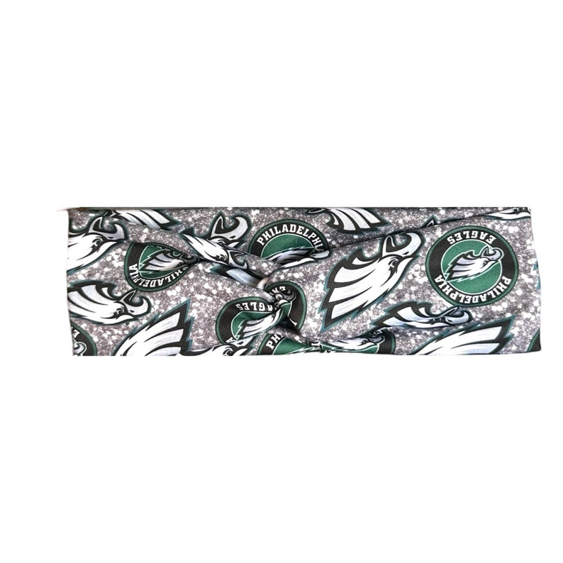 May include: A silver and green glitter headband with a repeating pattern of the Philadelphia Eagles logo. The logo features a green eagle head with the words "Philadelphia Eagles" in a circle around it.