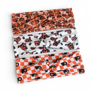 May include: Three fabric headbands with different patterns. The top headband is peach-colored with a repeating design of a cartoon dog. The middle headband is white with a repeating design of a hockey player. The bottom headband is orange and white checkered with a logo.