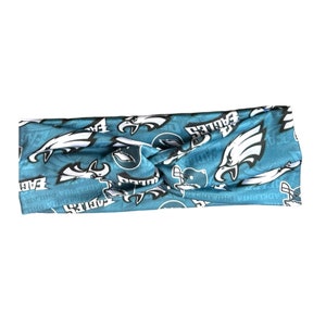 May include: A blue and white headband with a repeated pattern of the Philadelphia Eagles logo. The logo features a white eagle with a black beak and black eyes, and the word "Eagles" in black letters.