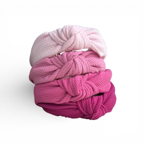 May include: A stack of four knotted headbands in shades of pink, from light to deep fuchsia. The headbands have a textured fabric and a knotted design at the top.