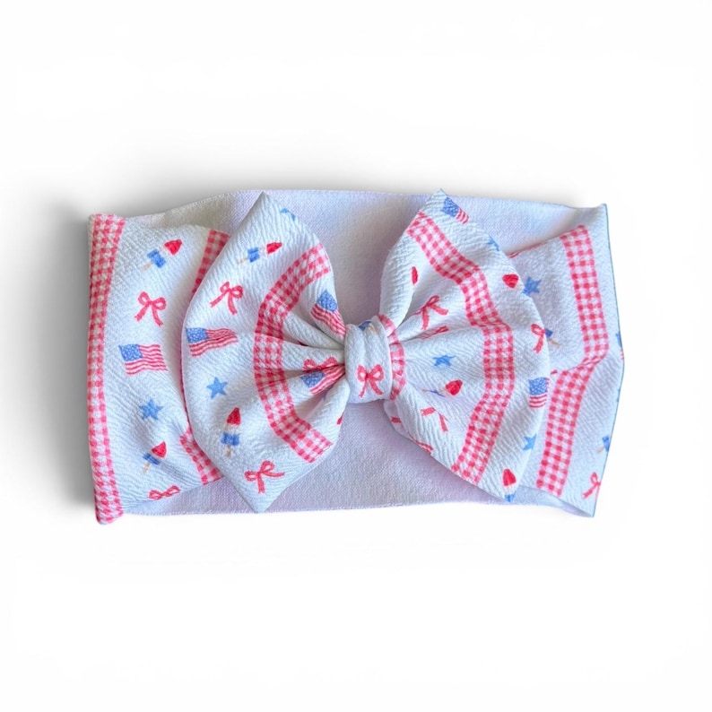 May include: White baby headband with a large bow featuring a patriotic design. The bow has a white background with red and blue accents, including stars, flags, and bows. The edges of the bow are trimmed with a red and white gingham pattern.