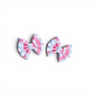 May include: Two fabric hair bows. Each bow features a white background with a pattern of red gingham, blue stars, and small American flags. The bows are tied in the center with a knot.