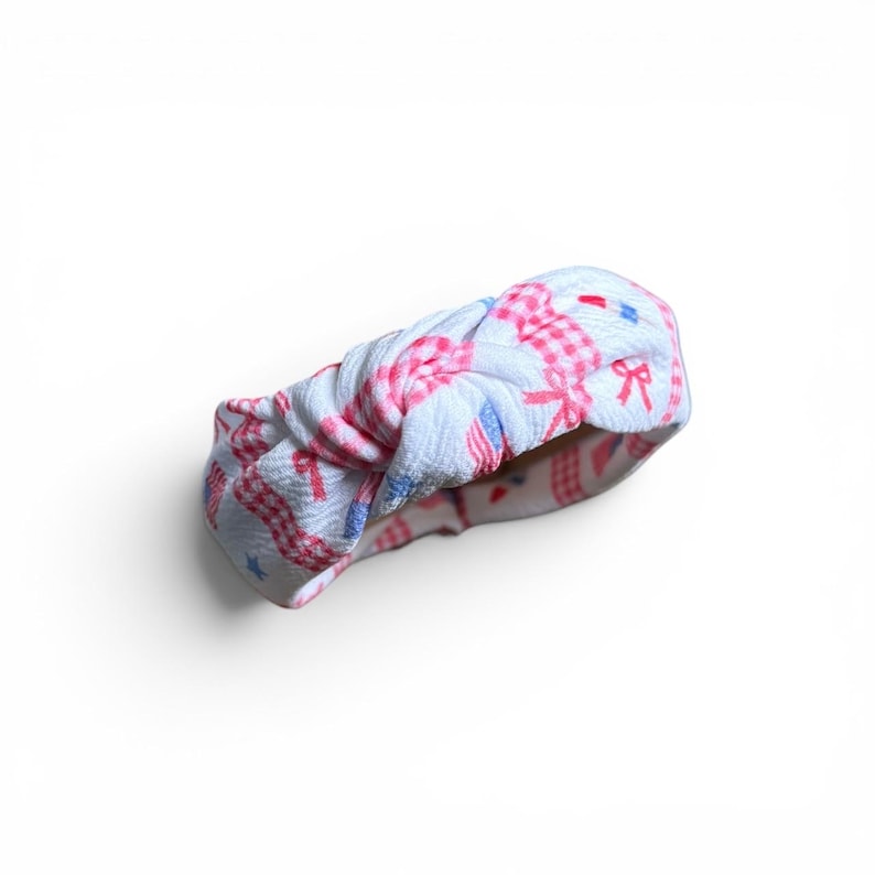 May include: White and pink patterned headband with a knotted design. The fabric features a gingham pattern with red bows, blue stars, and red and blue accents. This accessory is perfect for adding a touch of style to any outfit.