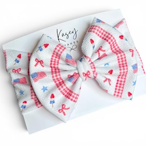 May include: A white baby headband with a large bow featuring a patriotic design. The bow has a gingham pattern in red and white, with scattered images of American flags, bows, stars, and popsicles. The headband is on a white card with the text "Kasey Makes".
