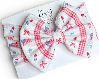 May include: A white baby headband with a large bow featuring a patriotic design. The bow has a gingham pattern in red and white, with scattered images of American flags, bows, stars, and popsicles. The headband is on a white card with the text "Kasey Makes".