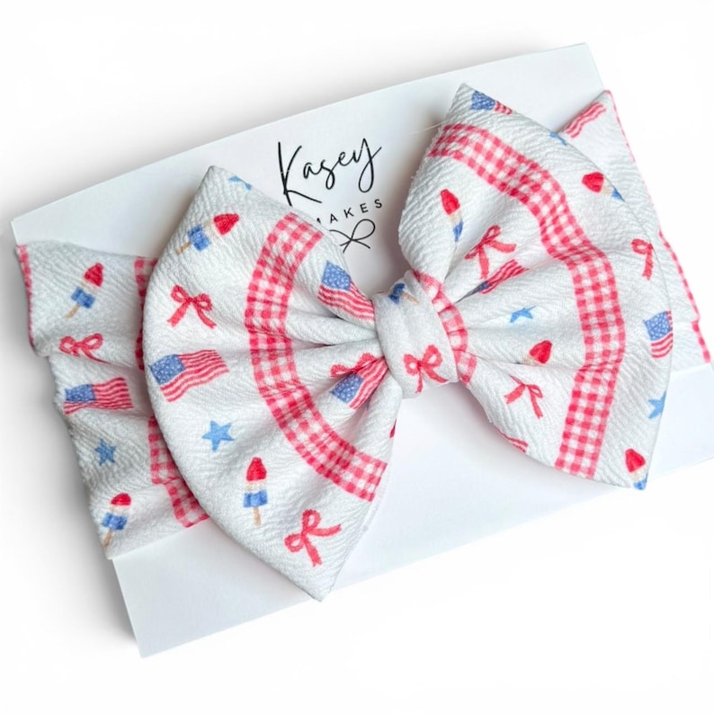 May include: A white baby headband with a large bow featuring a patriotic design. The bow has a gingham pattern in red and white, with scattered images of American flags, bows, stars, and popsicles. The headband is on a white card with the text "Kasey Makes".