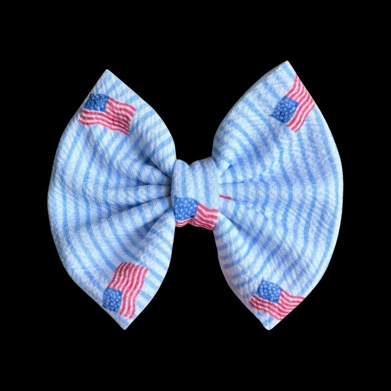 May include: A light blue bow with a striped pattern and small American flags. The bow is made of a soft fabric and has a knot in the center. The bow is a patriotic accessory.