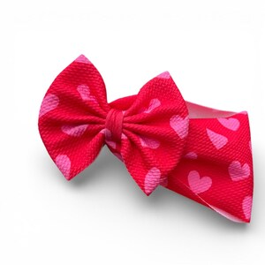 May include: A red baby headband with a large bow featuring a pink heart pattern. The headband is made of a soft, textured fabric and is designed to be comfortable for infants and toddlers.