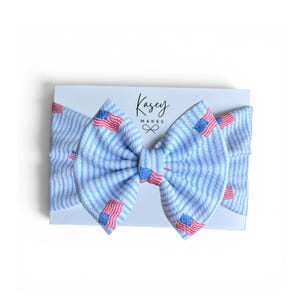 May include: A blue and white striped headband with red, white, and blue American flags. The headband is on a white card with the text "Kasey Makes". The bow is centered on the card.