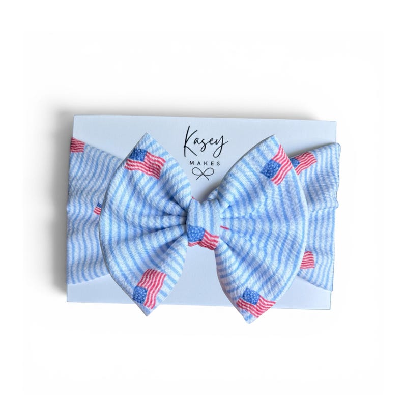 May include: A blue and white striped headband with red, white, and blue American flags. The headband is on a white card with the text "Kasey Makes". The bow is centered on the card.
